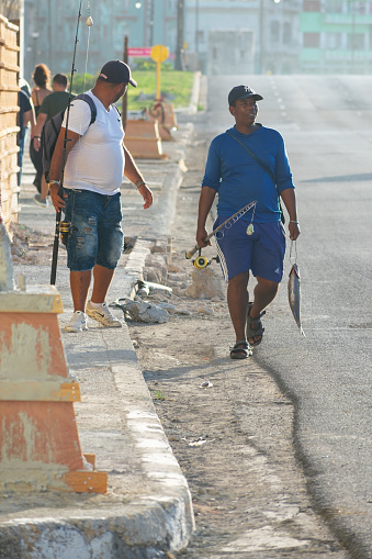 Havana, Cuba - September 16, 2022: Two fishermen walking on a sidewalk in a city street. The men hold a fishing rod with the reel in their right hands, and one has a fish in his left hand.