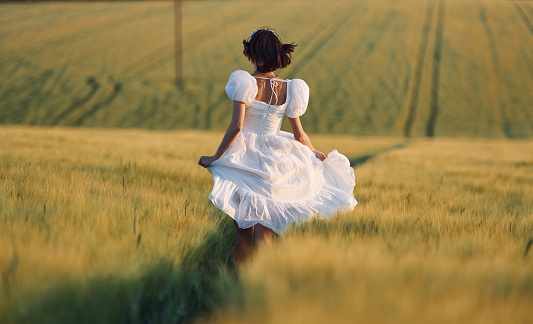 Rear view. Beautiful young bride in white dress is on the agricultural field at sunny day.