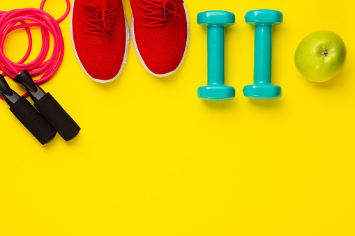 Fitness and sport concept. Jump rope, green apple, dumbbells and red sneakers on bright yellow background. Free space.