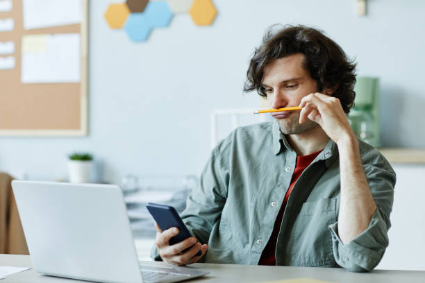 jeune homme ennuyé au bureau en utilisant le téléphone - lanterne photos et images de collection