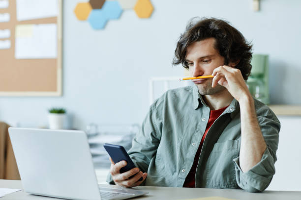 Bored Young Man in Office Using Phone Portrait of Caucasian young man playing with pencil and using phone at workplace suffering from boredom and procrastination ADHD in Adults stock pictures, royalty-free photos & images