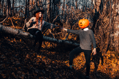 Witch woman sits on a birch tree and orders a man with a jack lantern on his head. Halloween scarecrow with a pumpkin reaches out with his hand to the enchantress. Concept of friendship and lovers.