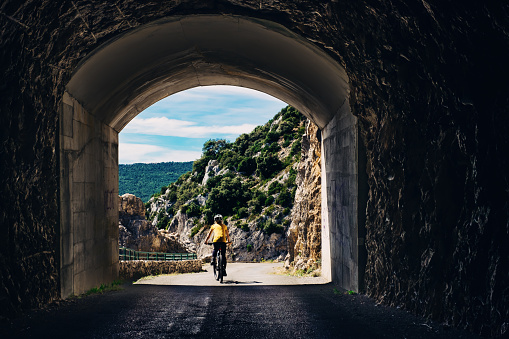 Mature female cyclist emerging from the road tunnel at the Belvedere des glacieres on the Route des Cretes at Verdon Gorge, Provence, France. Part of a series featuring all the main viewpoints.