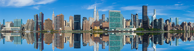 High Resolution Stitched Panorama of New York Skyline with UN Building (Headquarters of the United Nations), Chrysler Building, Empire State Building, Manhattan Upper East Side Residential and Office High-rises, FDR drive, Green Trees and Morning Blue Sky with Clouds All Reflected in water of East River. Canon EOS 6D (Full Frame censor) DSLR and Canon EF 85mm f/1.8 lens. 3.7:1 Image Aspect Ratio. This image is downsized to 50MP. Original image resolution was 121.2MP or 21295 x 5691 px.