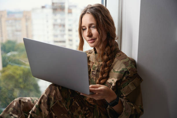 istock Woman in camouflage uniform sits on windowsill with a gadget 1434739166