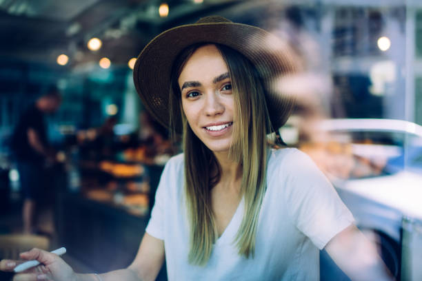 Trough glass view smiling young woman in tropical hat and white shirt looking at camera while sitting in cafe on blurred background