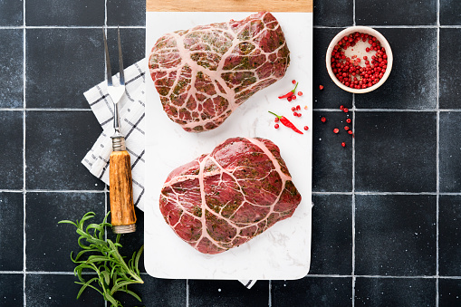 Steaks Raw. Raw cowboy steak, marbled beef meat with spices rosemary and pepper on white marble slate and grey table background. Top view. Mock up.