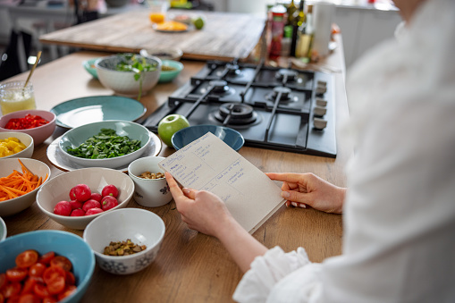 Closeup of the hands of a young Caucasian woman holding a paper with a list of ingredients for the preparation of a healthy salad. Her arms are leaning on a wooden table and in front of her there are many bowls with fresh chopped vegetables.