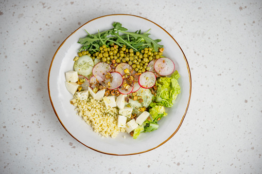 Aerial view of a green healthy salad in a white ceramic plate with arugula, green peas, cucumber and raddish slices, couscous, cheese pieces and pistachios as a garnish.