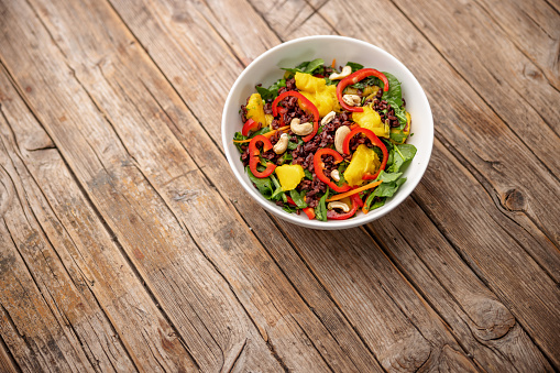 Aerial view of a colorful and healthy salad bowl with baby spinach, mango pieces, red bell pepper slices, dried cranberries and cashews. The salad bowl is placed over an old wooden table with cracks.