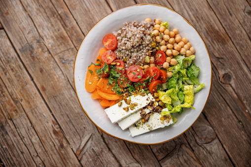 Aerial view of a colorful healthy salad containing sliced carrots, buckwheat, chickpeas, sliced lettuce, sliced cherry tomatoes, white cheese slices, garnished with chopped herbs and pistachio pieces. The salad is served on a white ceramic plate over a wooden table surface.