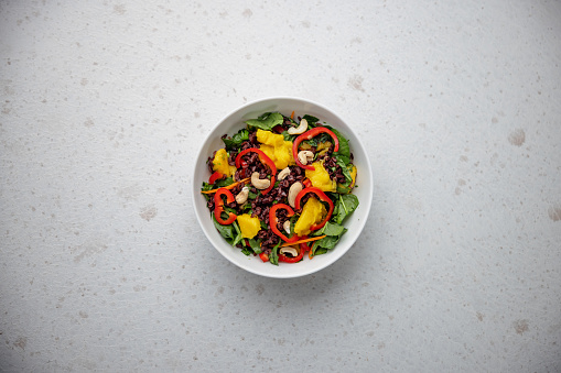 Aerial view of a colorful and healthy salad bowl containing raw baby spinachs, mango slices, red bell pepper slices, dried cranberries and cashews. The bowl is placed in the middle over a white granite kitchen surface.