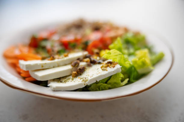 Close-Up of the white cheese of a vegetarian salad with some pistachio pieces on the top. The salad is served over a white ceramic plate with brown edge. The background of the picture is blurry-