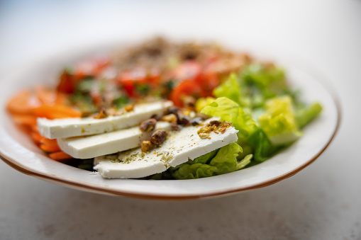 Close-Up of the white cheese of a vegetarian salad with some pistachio pieces on the top. The salad is served over a white ceramic plate with brown edge. The background of the picture is blurry-