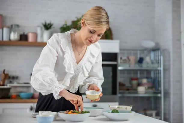 Portrait of a young caucasian woman adding ingredients to a colorful salad at the kitchen. She has a light smile and is holding a small bowl on her left hand. The background is a defocused modern kitchen. She is formally dressed and her hair is tied.