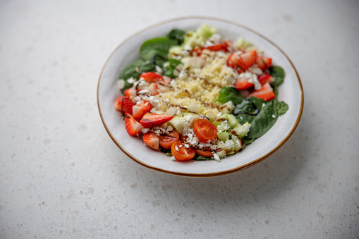 Closeup of a fresh colorful salad with blurry background. The salad contains sliced strawberries, raw spinach, sliced cherry tomatoes garnished with grated cheese and sliced almonds on the top.