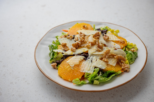 Side view of a white ceramic plate with a healthy colorful salad that contains lettuce, black beans, orange slices, grated cheese, walnuts and dressing. The salad plate is placed on a white granite surface.