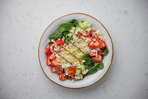 Side view of a colorful and healthy salad served on a white ceramic plate placed over a white granite surface. The salad contains spinach, strawberries, cherry tomatoes and garnished with sliced almonds, grated cheese and balsamic vinaigrette. The plate is placed on the bottom of a white granite surface