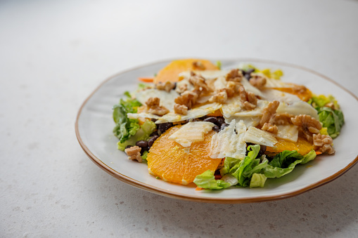 Closeup of a white ceramic plate with a colorful fresh salad that contains lettuce, black beans, orange slices, grated cheese, walnuts and dressing on the top. The salad plate is placed on a white granite surface.