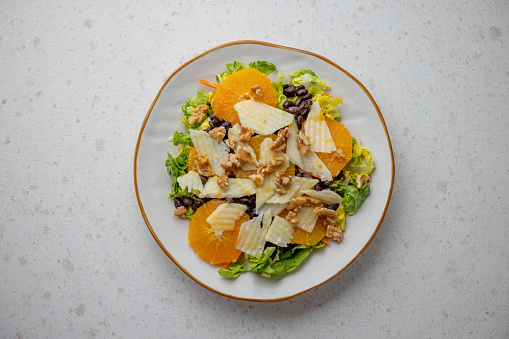 Aerial view of a white ceramic plate containing a colorful healthy salad. The salad has fresh lettuce, orange slices and black beans. It is garnished with cheese pieces, walnuts pieces and yellow dressing on the top.