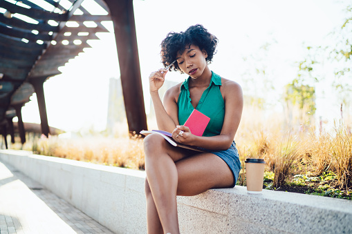 Pretty african american woman with curly hair reading interesting boor recreating on sunny summer day in city park,Pensive dark skinned female student concentrated on literature learning outdoors