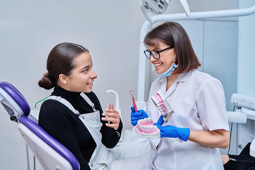 Teenage female sitting in dental chair at dentist checkup, doctor with dental jaw model and toothbrush telling and showing teenager dental care. Adolescence hygiene treatment dental health care