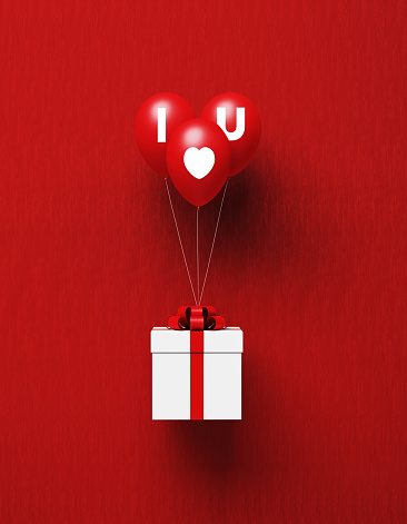 White gift box tied with red ribbon is about to be carried away by I Love You written red balloons on red background. Vertical composition with copy space, Great use for gift concepts.