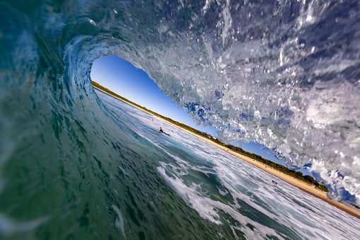 A closeup of a wave tunnel in the ocean with a person surfing and the beach in the background.