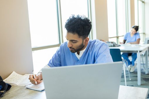 The young adult medical student takes notes while listening to an online podcast on his laptop.