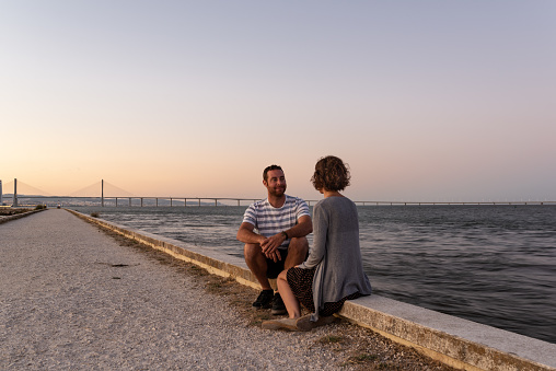 Man and woman couple enjoying a purple sunset at the riverbank of Tejo in Lisbon, Portugal.
