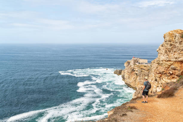 istock Standing man  with a backpack at the edge of a cliff while he is looking at Forte de Sao Miguel in Nazare, Portugal. 1434757282