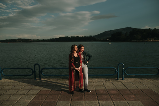 Full length portrait of two young women standing in a promenade by the sea, with their faces lit by the sun.