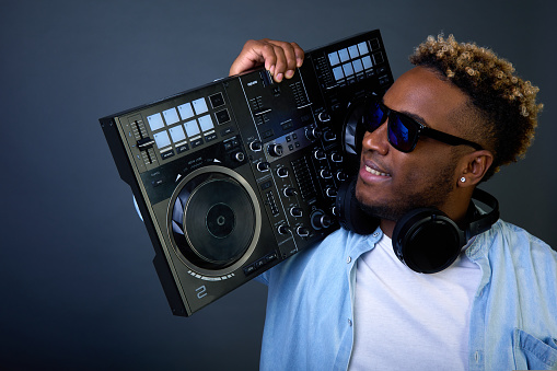 Cool guy with dark glasses on shoulder holds DJ console and carries club. Dark skinned DJ in headphones and glasses, in fashionable clothes is in hurry concert please youth in club with music track