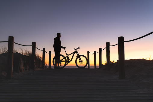 Silhouette of a man with his bike looking at a beautiful sunset at the beach.