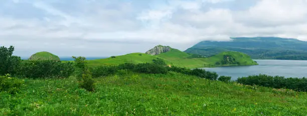 natural landscape of Kunashir island with grassy hills, volcanic rocks, volcano in the clouds and a valley with a lagoon