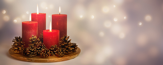 Vertical shot of three christmas candles on wood table. The candles are of differen sizes and are burning with calm flames. The composition is decorated with a red ribbon and two red baubbles, various pine cones and green pine leaves. In the foreground are defocused christmas lights giving a warm mood to the image. Photo taken in studio with DSRL Canon EOS 5D Mk II 