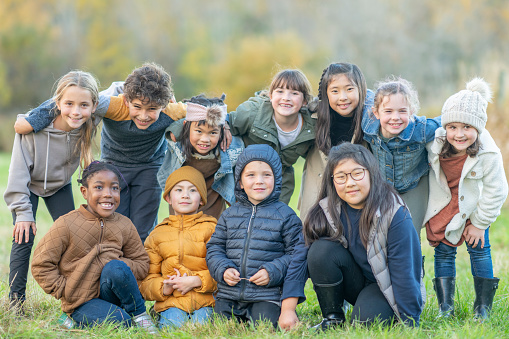 A large group of school aged children huddle in together, side-by-side, in two rows as they pose for a portrait.  They are each dressed warmly in fall layers as they stand with their arms around one another and smile.