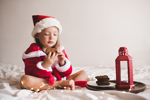 Cute child girl 3-4 year old wear red santa claus hat and bathrobe sit in bed with Christmas decor and glow lights at home. Kid eat chocolate biscuits. Winter holiday season