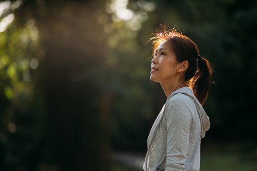 Side view of Asian Chinese woman jogging outside in sunny rainforest. She took a break to rest and looking away. The sun is shining in the background .