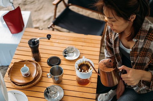 Top view of beautiful Asian woman pouring hot water from kettle to make drip coffee at campsite. Outdoor activity, adventure travel, or holiday vacation concept.