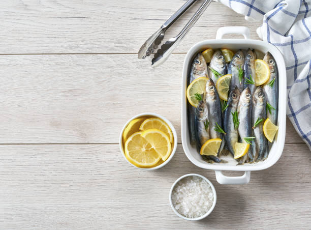 Marinated sardines with salt, herbs and lemon slices ready to cooking on a white table, copy space for text. Raw fish cooking and ingredients.