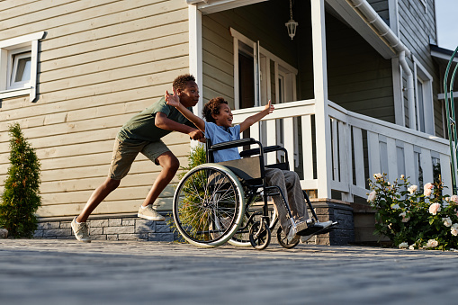 Side view portrait of black teenage boy pushing littlle brother in wheelchair while having fun together outdoors