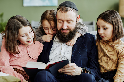 Portrait of orthodox Jewish man wearing kippah while reading book to three children