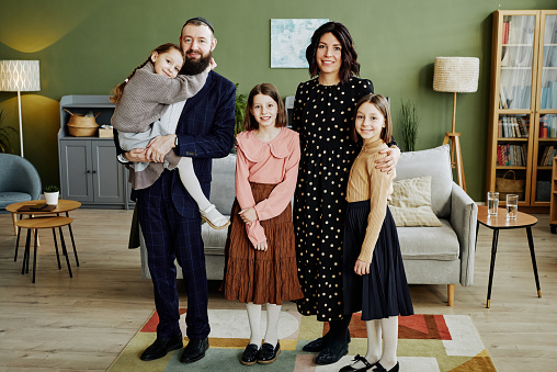 Full length portrait of happy jewish family standing in modern home and looking at camera with women and girls wearing dresses