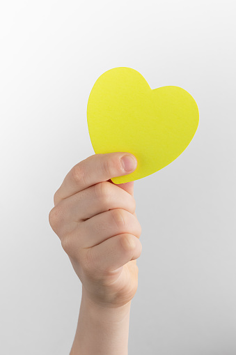 child hand holding yellow blank reminder or paper notes in the shape of a heart above a white and gray background, copy space