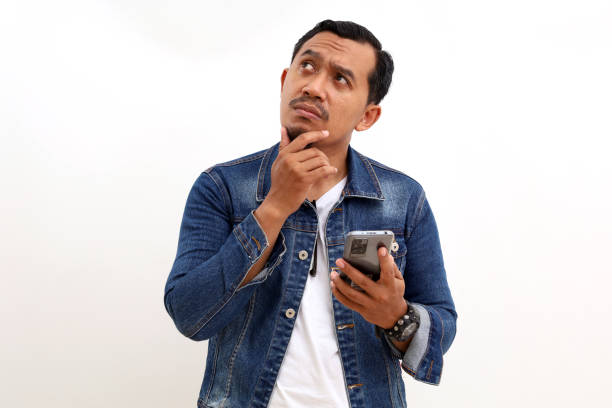 istock Thoughtful asian man standing while holding a cell phone. Isolated on white background 1434822175