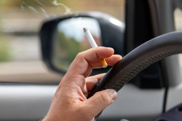 Close-up detail view of male hand driver holding cigarette while driving inside car on higway city street road on day time. Man person smoking inside vehicle. Low concentration attention danger stock photo