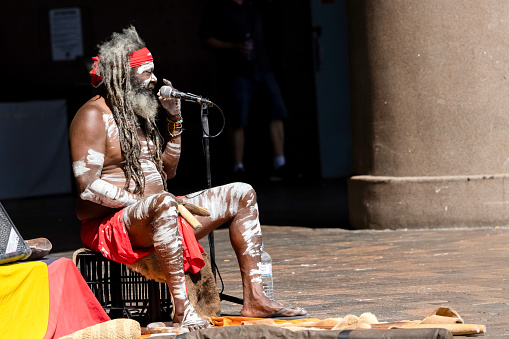 Sydney, Australia - October 16, 2022: Aboriginal male busker singing and performing with clap sticks at Circular Quay