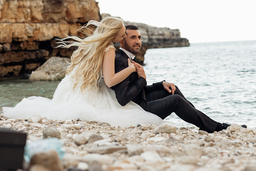 Side view of beautiful couple sitting on stones pebbles at shore near water. Young woman bride with long fair hair wearing long white wedding dress hugging kissing man groom. Wedding, honeymoon trip.