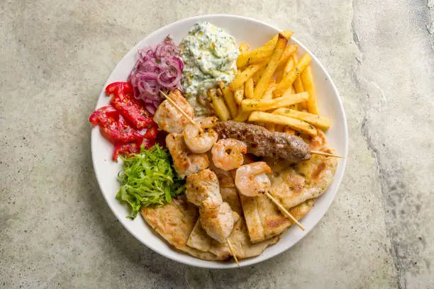 souvlaki with chicken, shrimp kebab and beef lula kebab with french fries, vegetables and pita on white plate on stone table top view